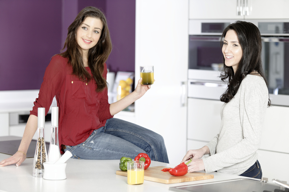 Two attractive young women preparing food in a white kitchen while talking.-1